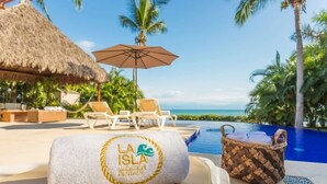 Zona de piscina frente al mar con sillones de playa, una sombrilla de paja y una toalla con logotipo.