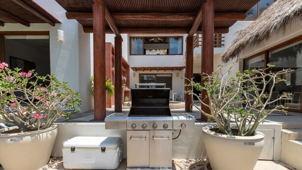 Un área de parrilla exterior con horno empotrado, rodeada de plantas en macetas, bajo una pérgola de madera.