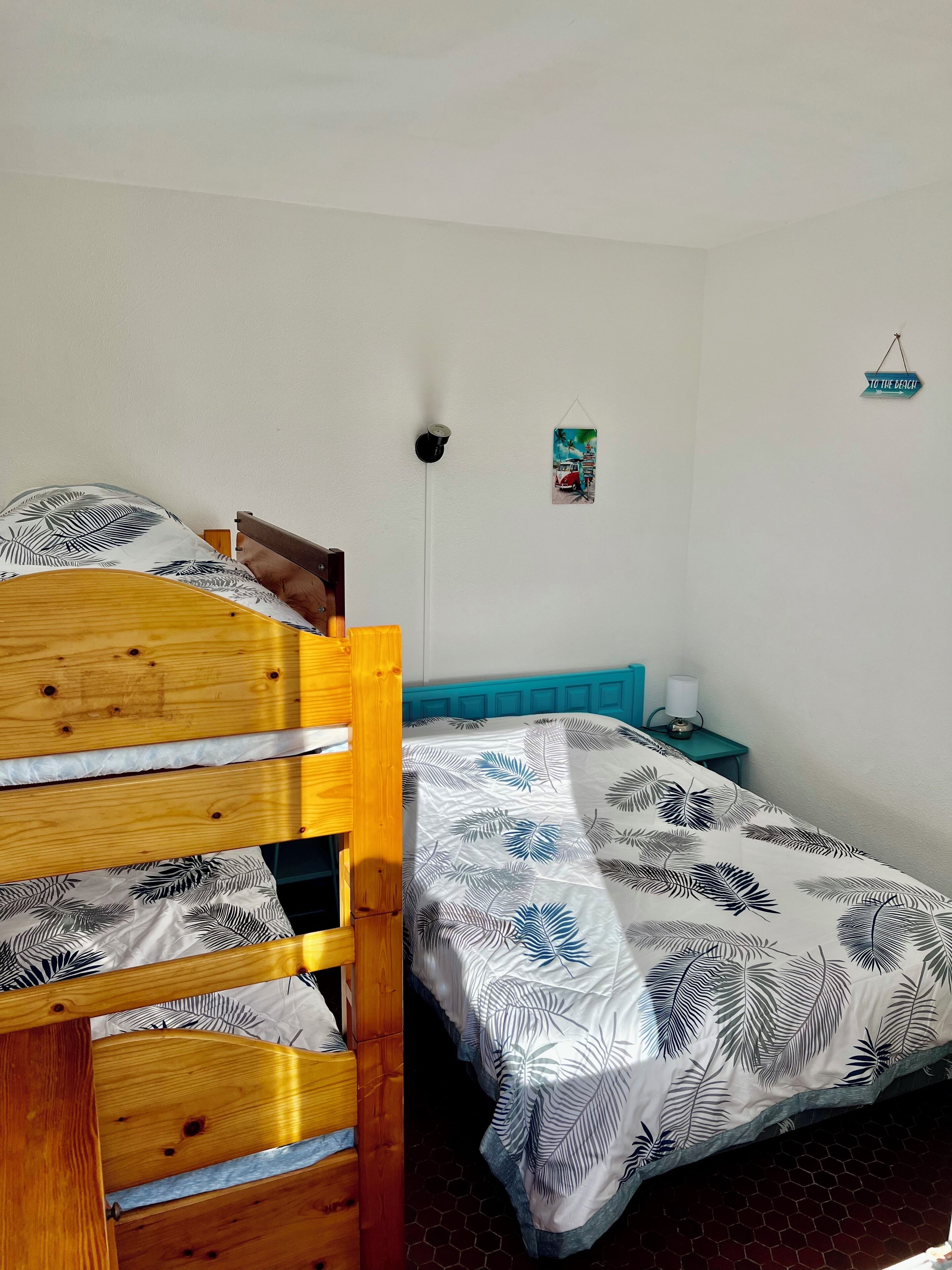 Un dormitorio con estructura de cama de madera, cama con cubrecama estampado con hojas, mesita de noche color turquesa con una lámpara y un marco de foto en la pared.