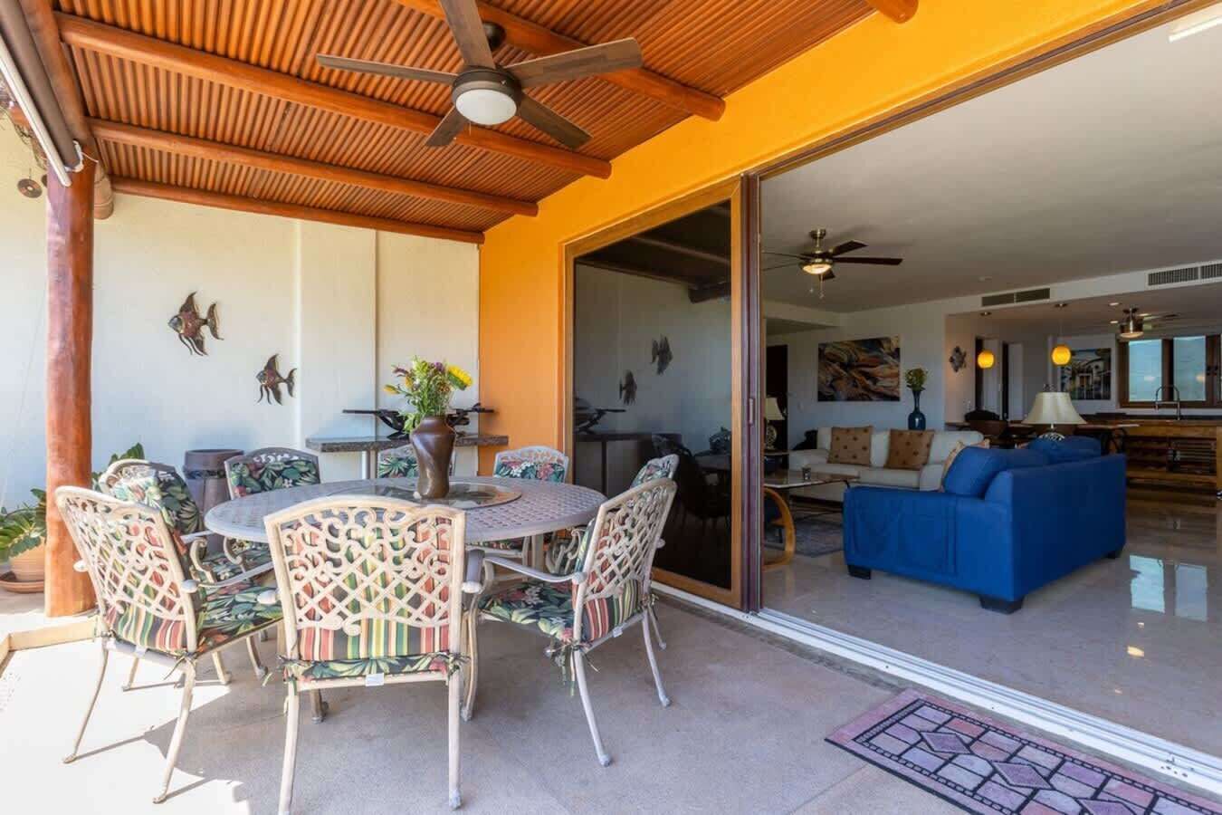 Un patio exterior con una mesa redonda y sillas, un ventilador de techo y un ventanal corredero que da a un salón.