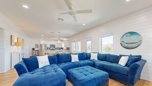 A spacious living room with a large blue sectional sofa and ottoman, a ceiling fan, and a kitchen in the background. - Private Pool, Palapa, Golf Cart Access (Port Aransas)