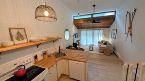 A modern kitchen with wooden countertops, a built-in oven, and a red kettle. There is a dining area with a sofa and a ceiling fan.