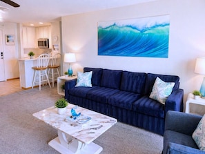 Een woonkamer met een donkerblauwe bank, een witte salontafel met een blauw decoratief object, een keuken met witte kasten en een magnetron, en een groot schilderij van een golf op de muur.