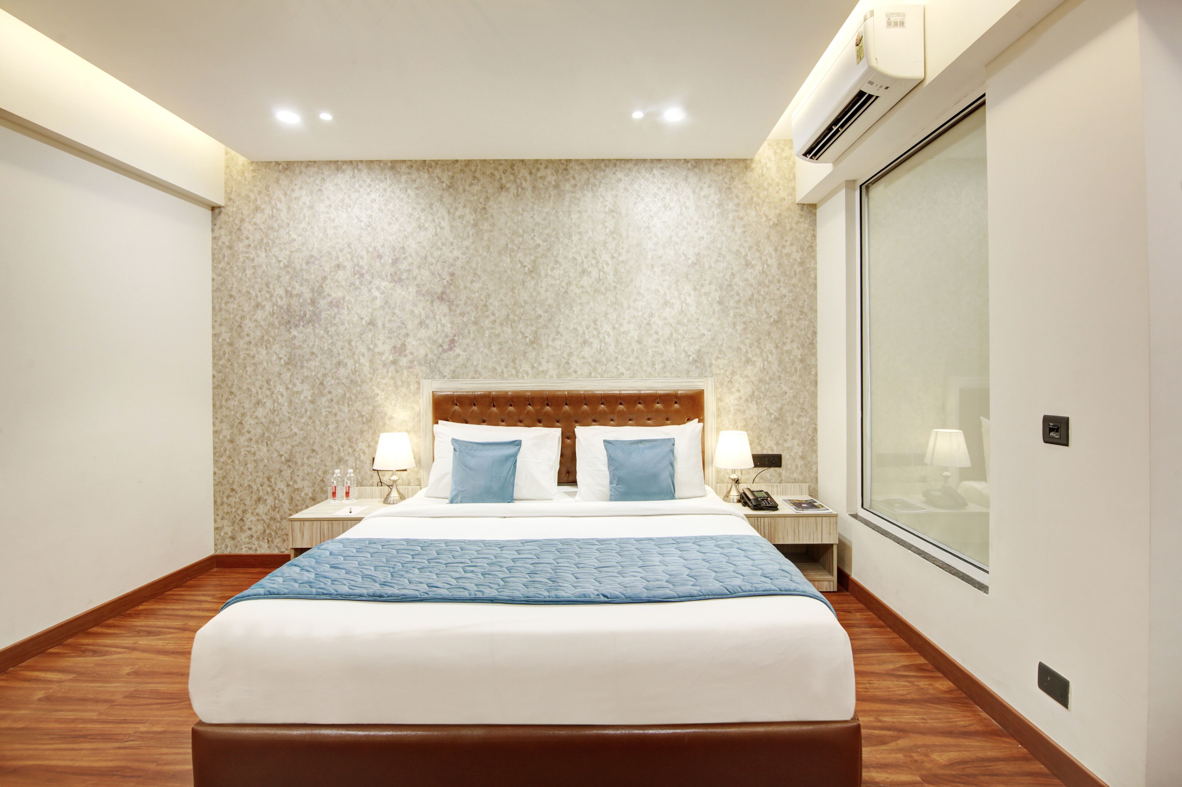 A hotel room with a large bed, two bedside tables with lamps, a wall-mounted air conditioner, and a large window.