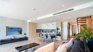 A modern living room with a sofa, a coffee table, and a TV mounted on the wall. There is a dining area with a table and chairs, and a kitchen in the background. - SeaView 3BR Duplex BluePoint 88 Patong (Patong)