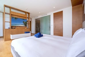 A modern hotel room with a large bed, a TV on a wooden shelf, a walk-in closet, and a glass door leading to a bathroom. - SeaView 3BR Duplex BluePoint 88 Patong (Patong)