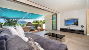 A modern living room with a large sofa, a glass coffee table, and a flat-screen TV mounted on the wall. There is a balcony with a blue awning and a view of the outdoors. - SeaView 3BR Duplex BluePoint 88 Patong (Patong)
