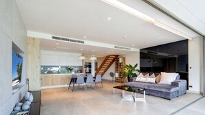 A spacious living area with a grey sofa, a coffee table, and a dining table with chairs. There is a staircase leading to an upper level, and a large window providing a view of the outdoors. - SeaView 3BR Duplex BluePoint 88 Patong (Patong)