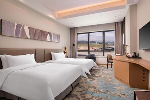 Ein Hotelzimmer mit zwei Betten, einem Schreibtisch, einem Stuhl, einem Fernseher und einem großen Fenster mit Blick auf einen Flughafen.