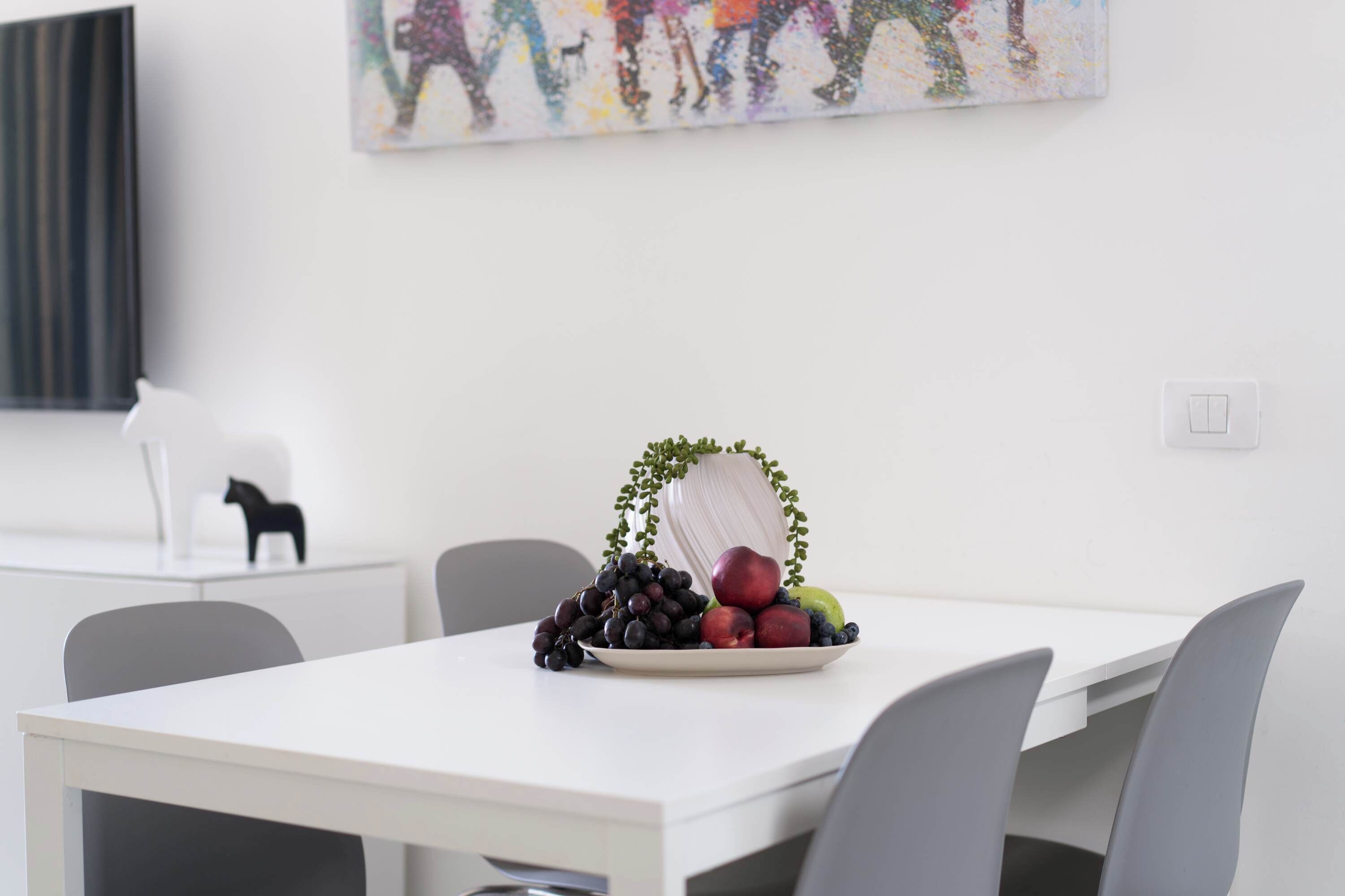 A modern dining area with a white table, grey chairs, and a decorative fruit arrangement.