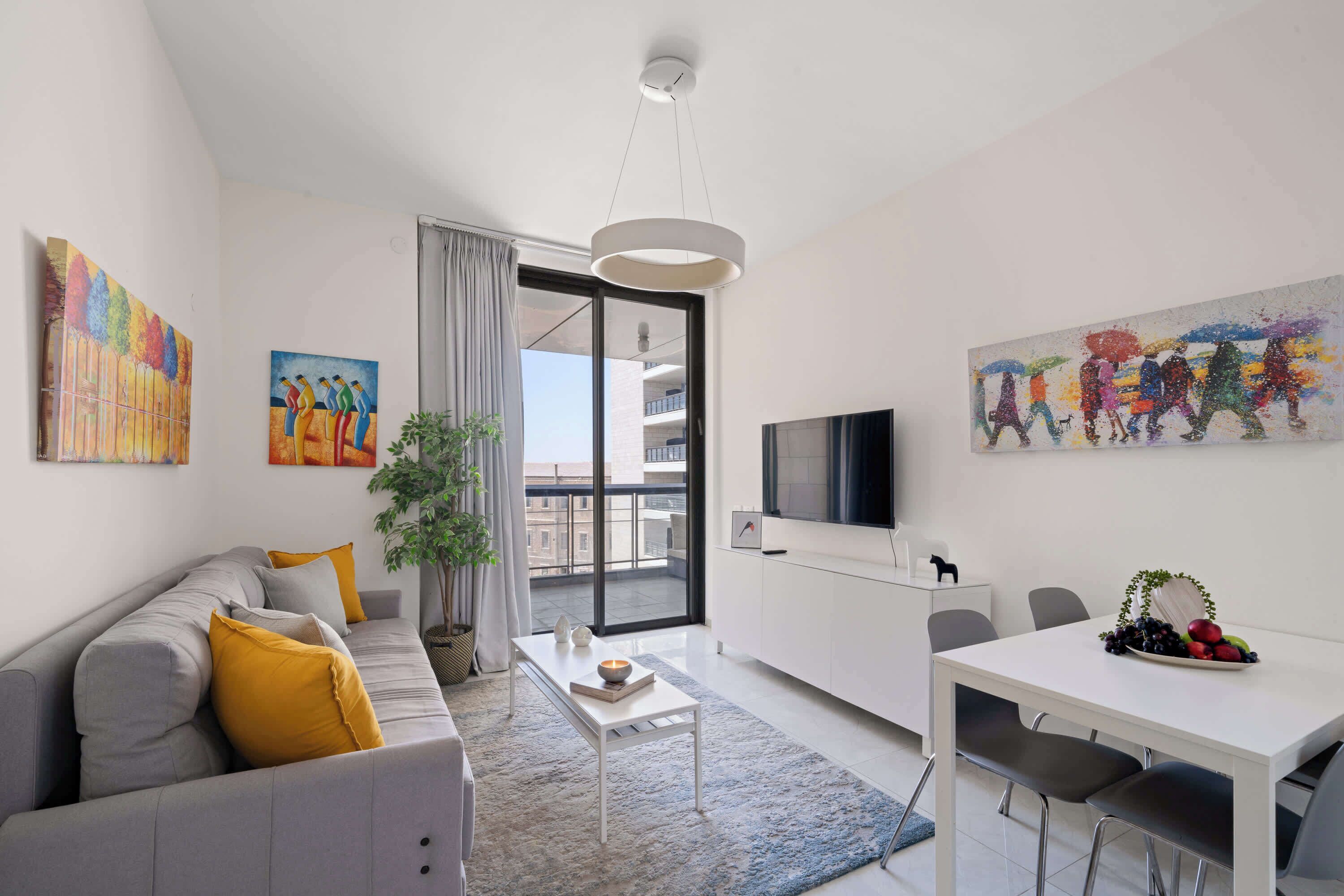 A modern living room with a grey sofa, a white coffee table, and a dining area with a white table and chairs. There are two paintings on the walls, a TV mounted on the wall, and a balcony visible through the door.