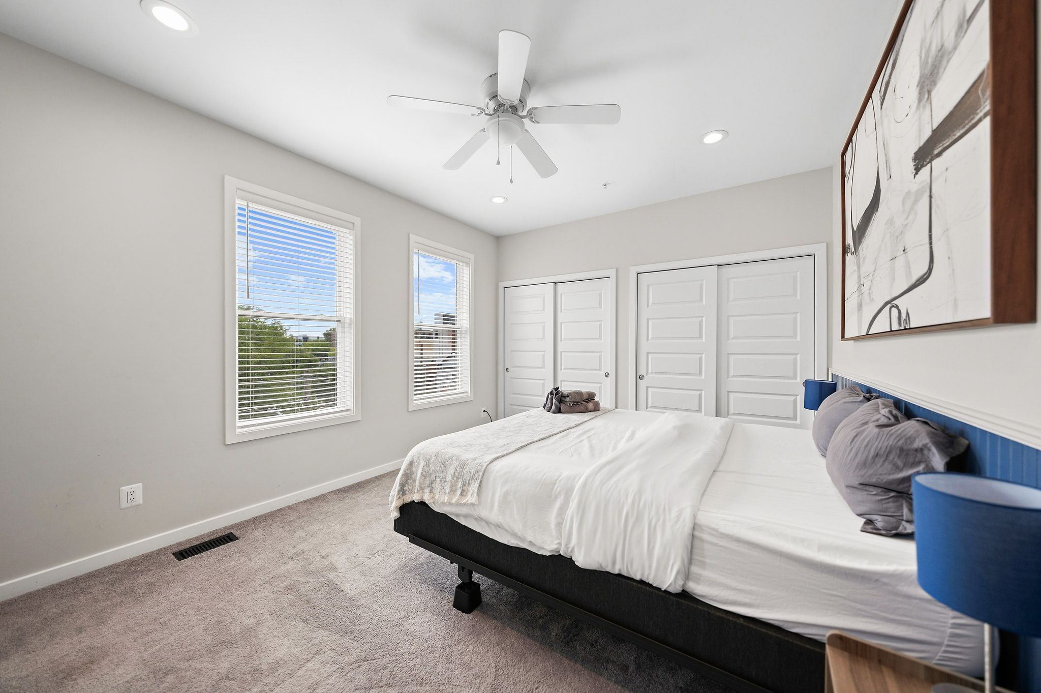A bedroom with a bed, a ceiling fan, two windows with blinds, a door, and a wall-mounted artwork.