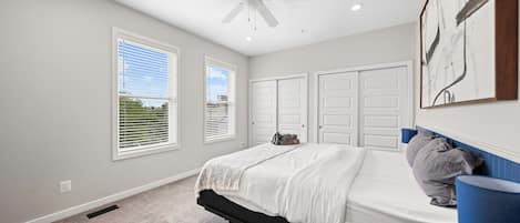 A bedroom with a bed, a ceiling fan, two windows with blinds, a door, and a wall-mounted artwork.