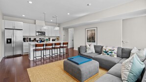 A modern living room with a grey sofa, a blue ottoman, and a kitchen area with white cabinets and a microwave.