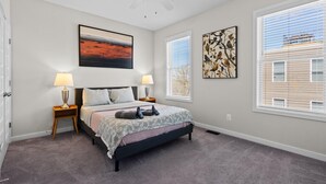 A bedroom with a bed, two bedside lamps, a ceiling fan, and two large windows with blinds.