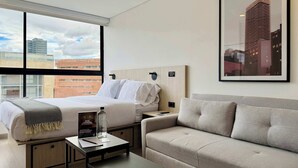 A hotel room with a bed, a sofa, a coffee table, and a large window offering a city view.