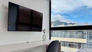 A modern room with a flat-screen TV mounted on the wall, a small white vase with decorative items, a grey chair, and a large window offering a view of buildings and mountains.