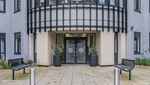 En bygningseingang med moderne design, med en buet facade og en glasdør. To plantekrukker flankerer indgangen, og foran er der to bænke.