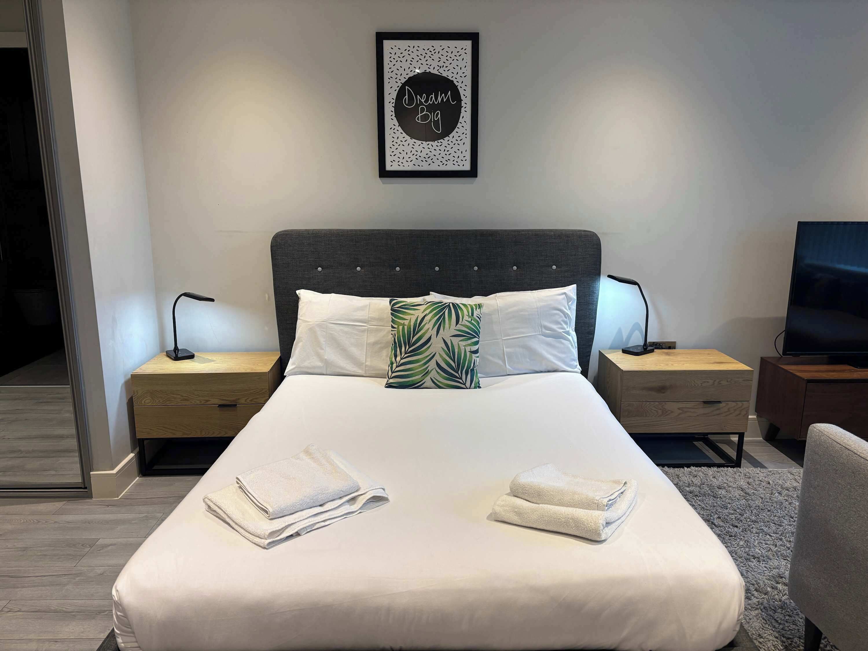 A neatly made bed with white linens and two folded towels, a dark upholstered headboard, a wall-mounted picture frame with text, bedside tables with lamps, and a television on a wooden stand.