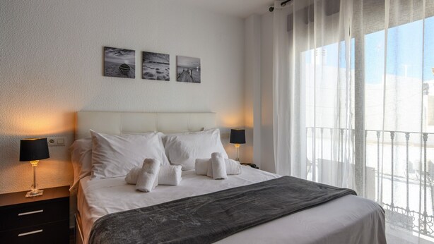 A hotel room with a large bed, two bedside tables with lamps, a view of the outdoors through sheer curtains, and two wall-mounted pictures.
