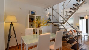 A modern living room with a glass dining table, white chairs, a sofa, and a staircase with metal railings.