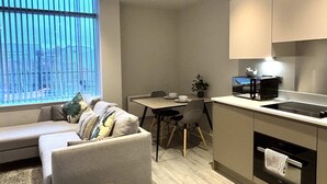 Ein gemütlicher Wohnbereich mit Sofa, Esstisch, Stühlen, einer kleinen Küchenzeile mit Mikrowelle und Backofen sowie einem Fenster mit Jalousien.