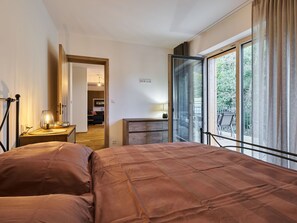 A bedroom with a large bed, a desk with a lamp, a sliding glass door to a balcony, and a view of greenery outside.