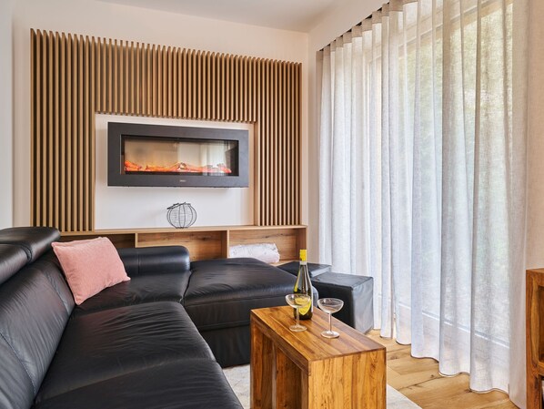 A modern living room with a black leather sofa, a wooden coffee table with a bottle and glasses, a flat-screen TV mounted on a wooden panel, and sheer curtains on a window.