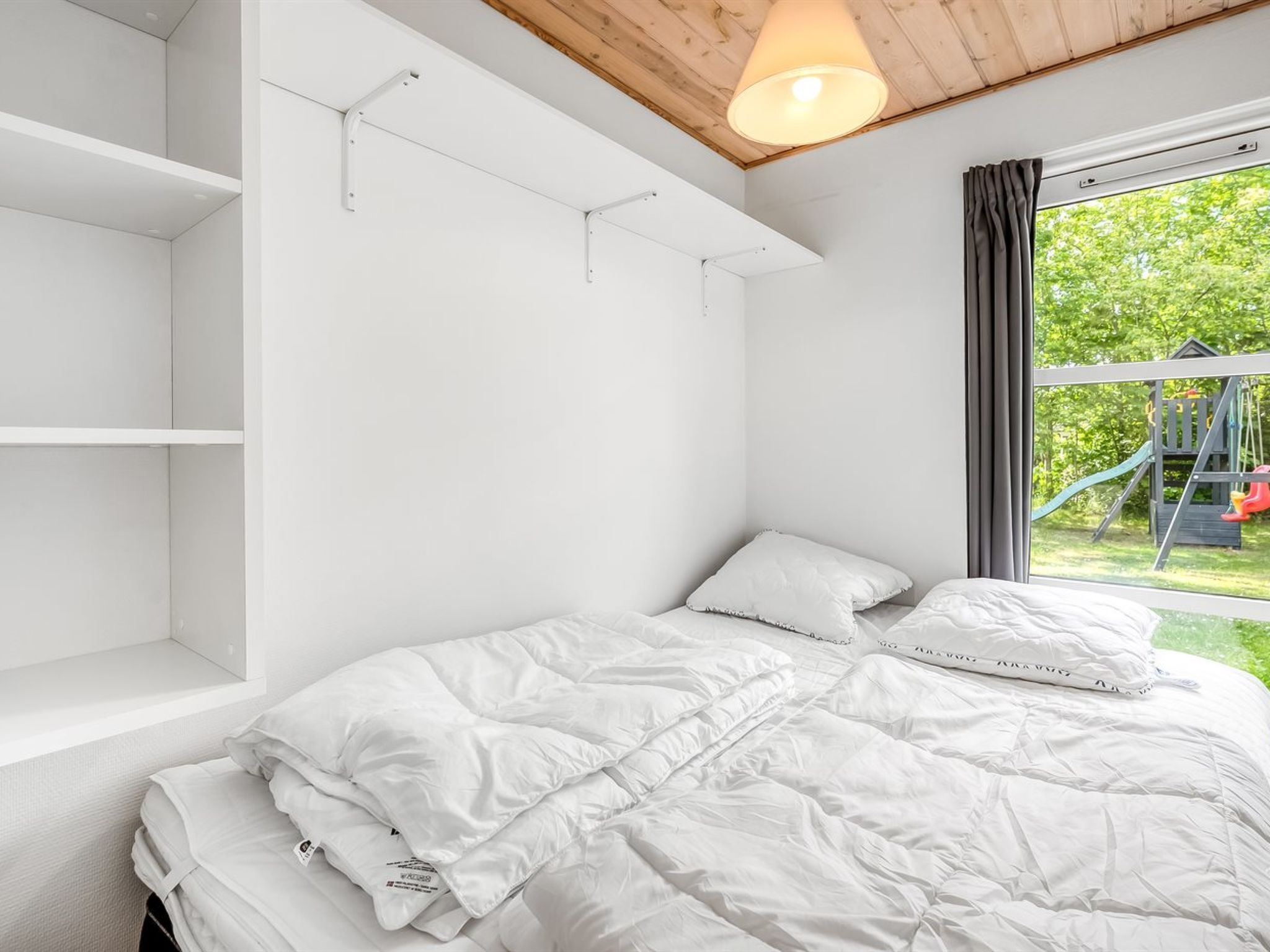 A bedroom with a bed, white bedding, a window with a view of a playground, and built-in shelves.
