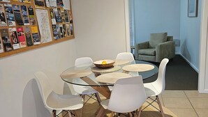 A dining area with a glass table surrounded by white chairs, a woven placemat, and a bulletin board with travel posters. - Mornington peninsula getaway (Frankston)