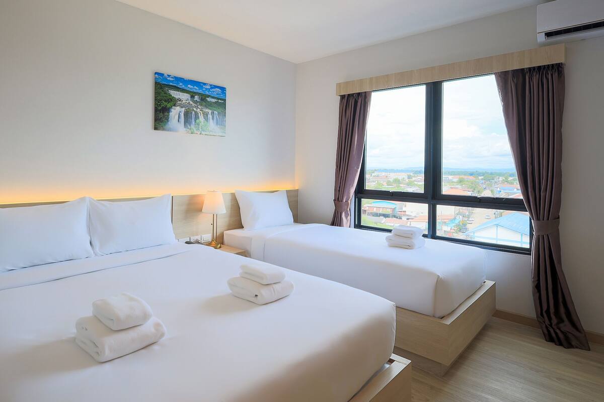 Ein Hotelzimmer mit zwei Betten, einem Bild an der Wand, einem Fenster mit Vorhängen und Blick auf die Stadt.