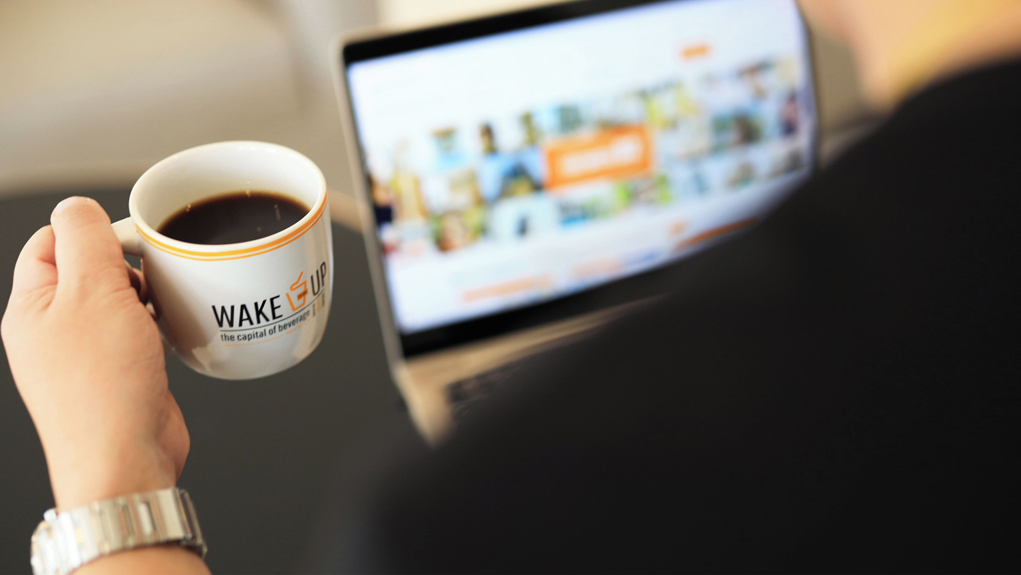 Una mano sosteniendo una taza de café con un logotipo, y una computadora portátil mostrando una imagen borrosa de un edificio.