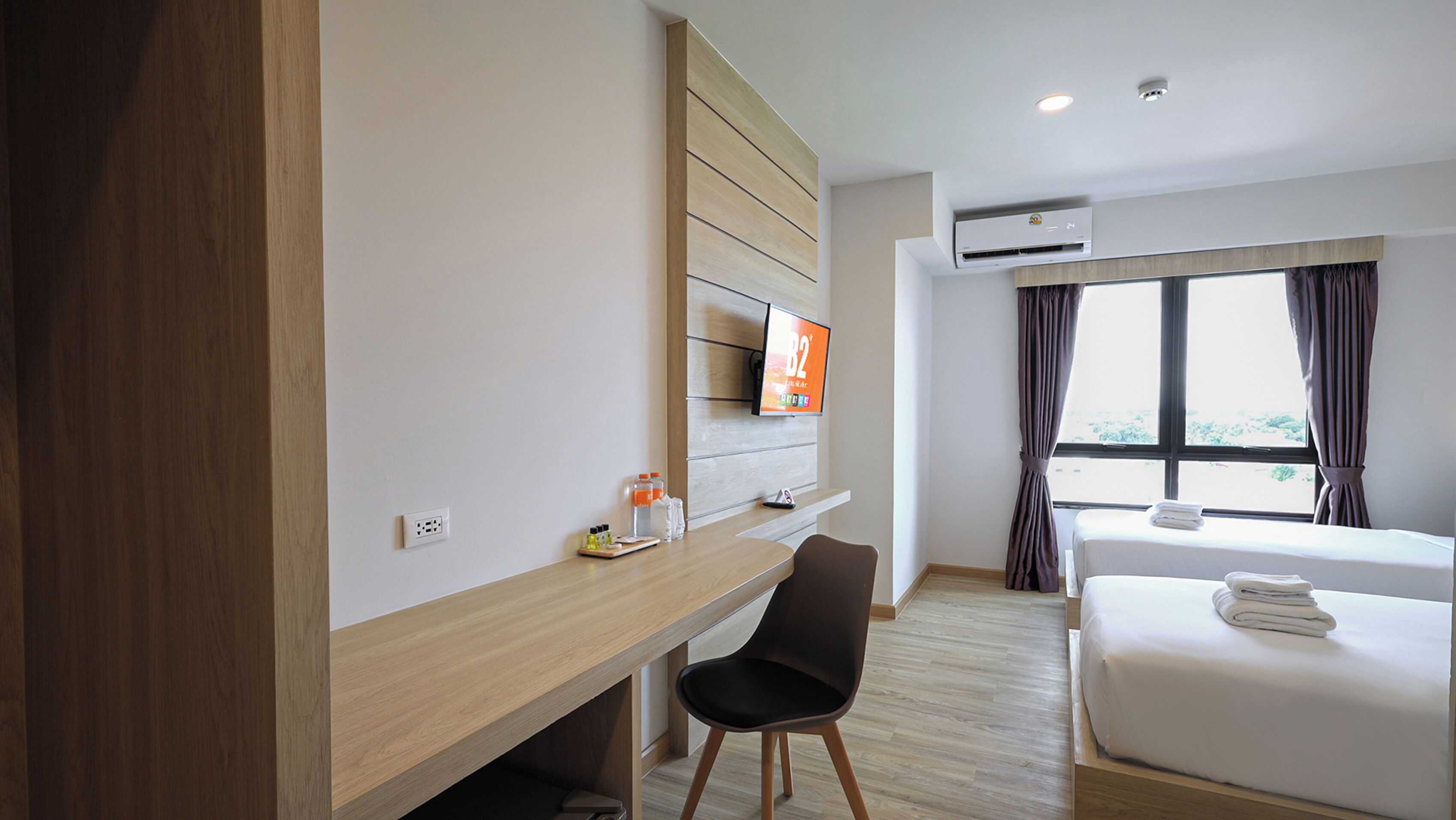 A hotel room with a wooden desk, a flat-screen TV, a bed, and a window with curtains.