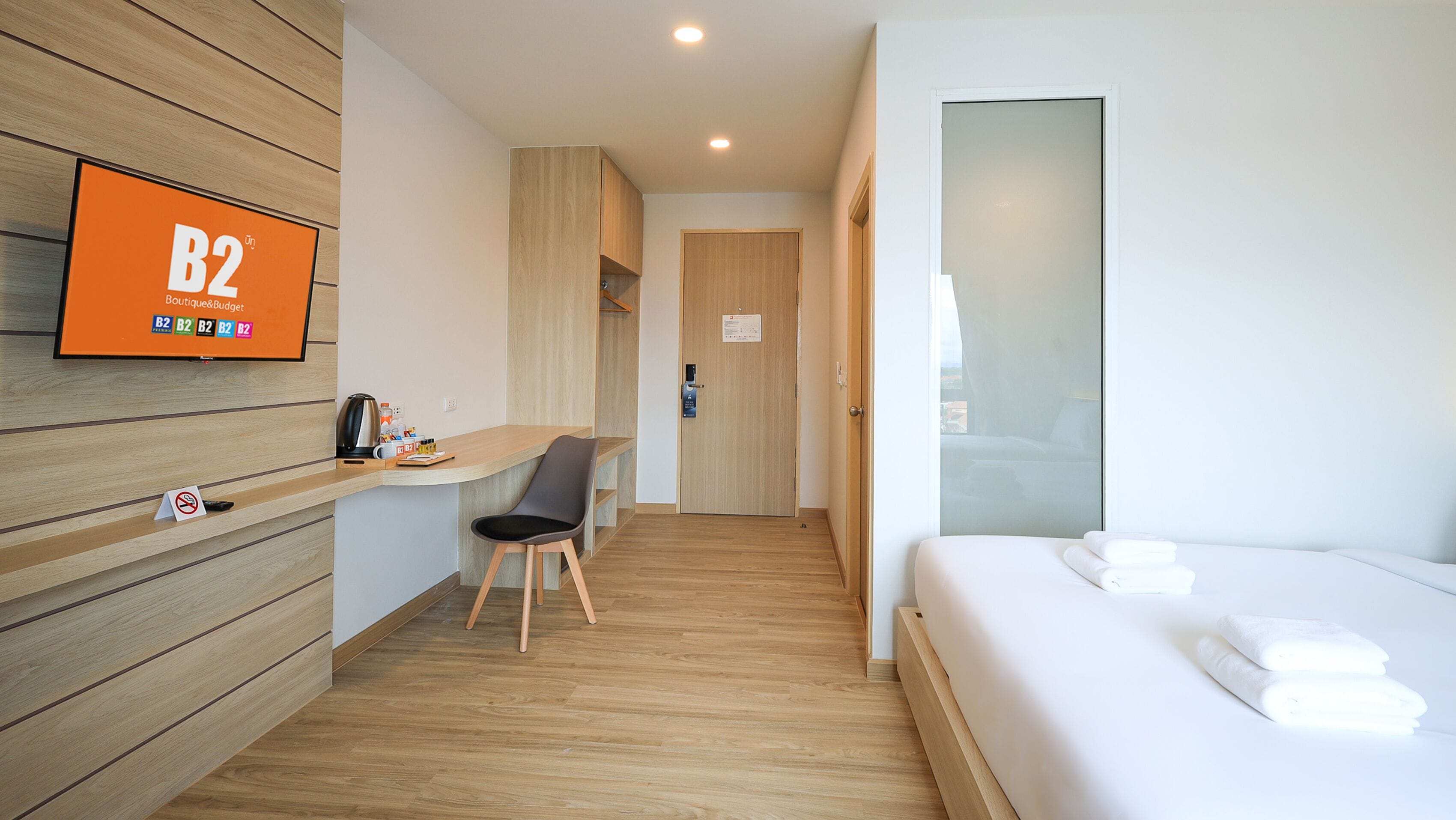 A hotel room with a bed, a desk, a chair, a TV mounted on the wall, and a door leading to the hallway.