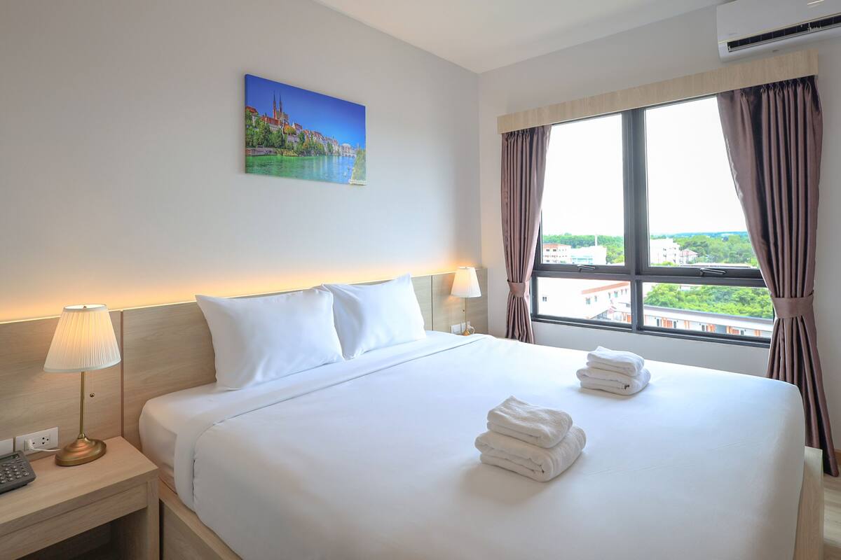 Ein Hotelzimmer mit einem großen Bett, zwei Nachttischlampen, einem Stadtbildgemälde und Blick auf Gebäude durch das Fenster.