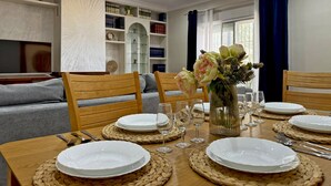 A dining area with a table set for six, surrounded by wooden chairs, a vase with flowers, and a bookshelf in the background. - Villa Victoria - Ole Solutions (Puerto de Santa María)
