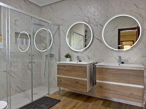 Ein modernes Badezimmer mit Duschkabine aus Glas, zwei Waschbecken mit Holzschränken und drei runden Spiegeln mit beleuchteten Rahmen.