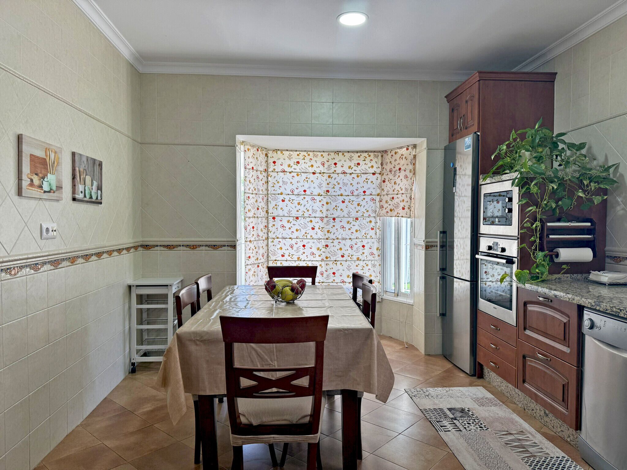 A kitchen with a dining table, chairs, a refrigerator, and a window with patterned curtains.