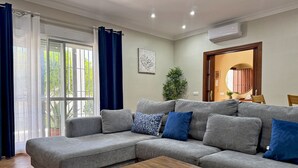 A living room with a grey sectional sofa, a wooden coffee table, a large window with curtains, and an air conditioning unit. - Villa Victoria - Ole Solutions (Puerto de Santa María)