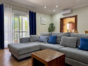 Una sala de estar con un sofá seccional gris, una mesa de centro de madera, un ventanal con cortinas y un equipo de aire acondicionado.