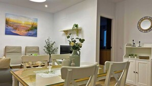 A modern dining area with a glass-top table, white chairs, a vase of flowers, and a painting of a sunset on the wall. - Apartamento Alborada - Ole Solutions (Sanlúcar de Barrameda)