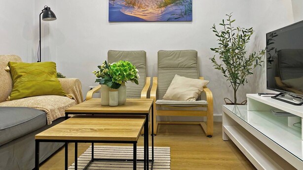 A modern living room with a sofa, chairs, a coffee table, a TV, and a painting of a sunset over water. - Apartamento Alborada - Ole Solutions (Sanlúcar de Barrameda)