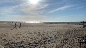 A beach with a clear sky and the sun shining. There are people walking and sitting on the sand, and a few boats in the water. - Apartamento Alborada - Ole Solutions (Sanlúcar de Barrameda)