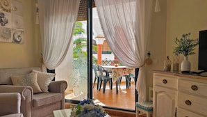 A living room with a sofa, a coffee table with a floral arrangement, a television on a cabinet, and a sliding glass door leading to an outdoor patio with a table and chairs. - Zahara Paradise - Ole Solutions (Zahara de los Atunes)