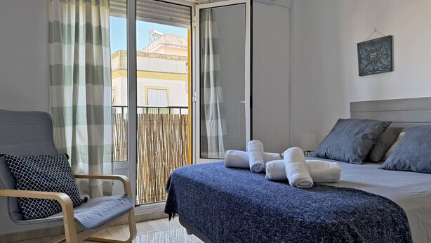 A hotel room with a bed, a chair, a balcony with curtains, and an air conditioning unit. - Apartamento La Paquera - Ole Solutions (Jerez de la Frontera)