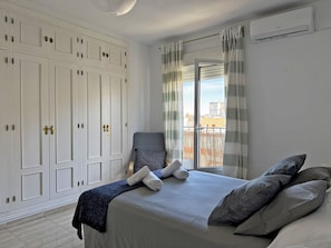 Un dormitorio con cama, una silla, un armario y vistas por la ventana.