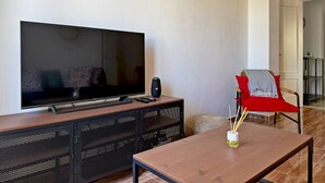 A living room with a flat-screen TV on a wooden cabinet, a red chair with a grey throw, and a decorative fan on the wall. - Apartamento La Paquera - Ole Solutions (Jerez de la Frontera)