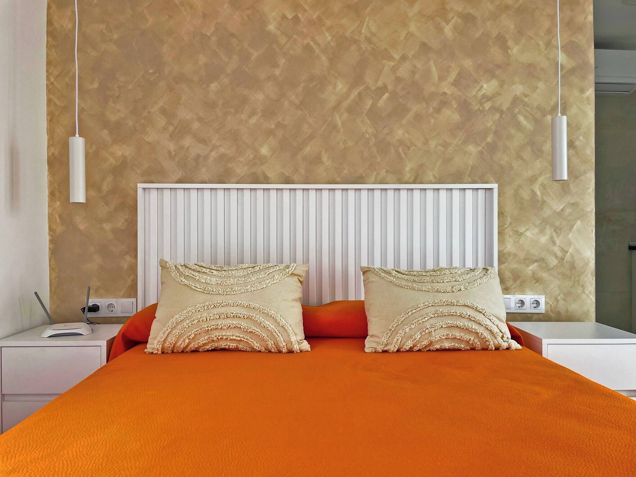 A modern bedroom with a large bed, two decorative pillows, a white bedside table, and a wall-mounted light fixture.