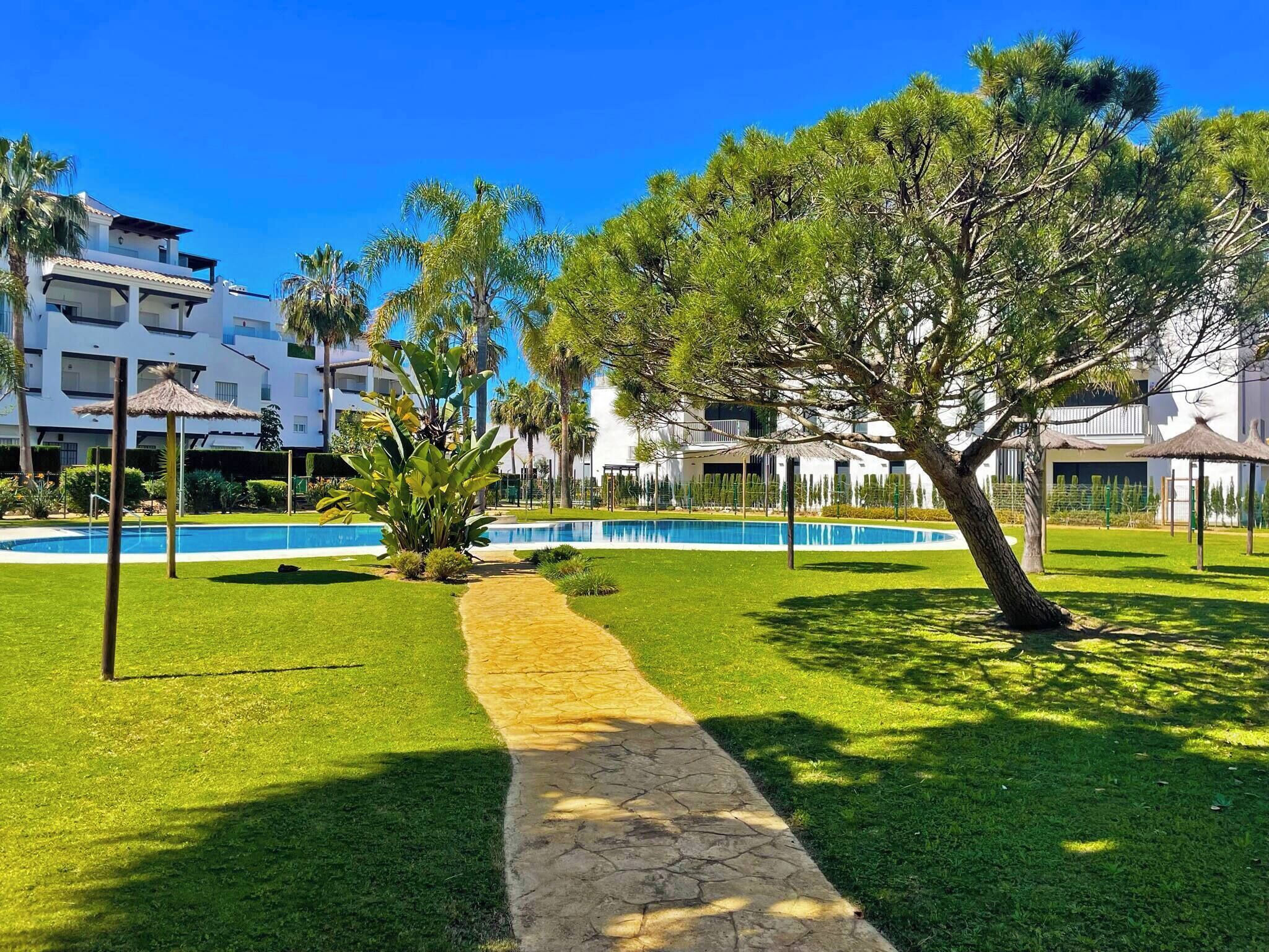 A pool area with a stone pathway, surrounded by well-maintained grass and trees, with modern buildings in the background.