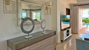 A modern living room with a large mirror, two wall sconces, a glass-top console table, a television, and a view of a pool and garden. - Villa Los Olivos - Ole Solutions (Arcos de la Frontera)